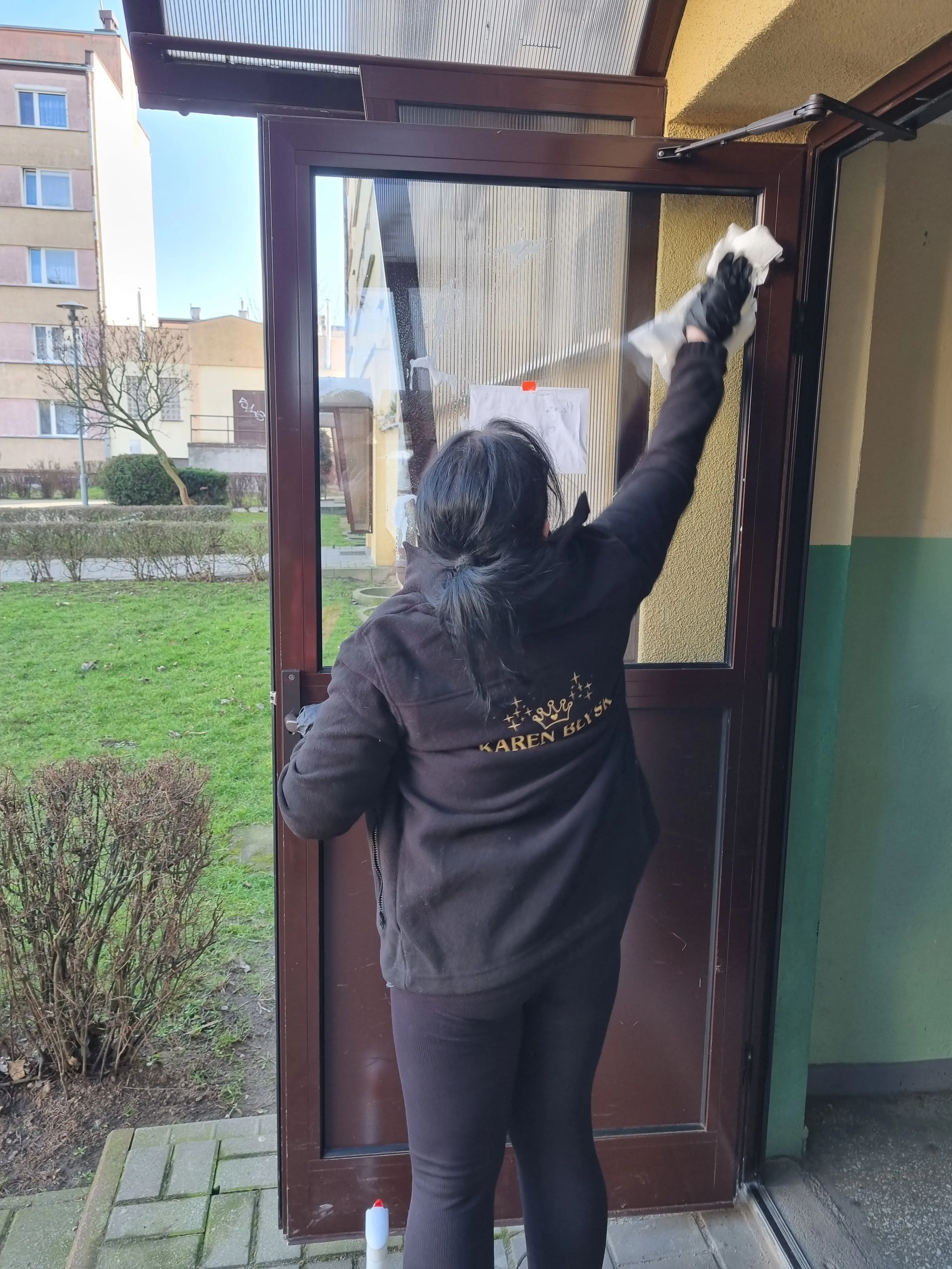 Sprzątaczka myjąca szybę w drzwiach do klatki schodowej – profesjonalne sprzątanie klatek schodowych przez Karen Błysk